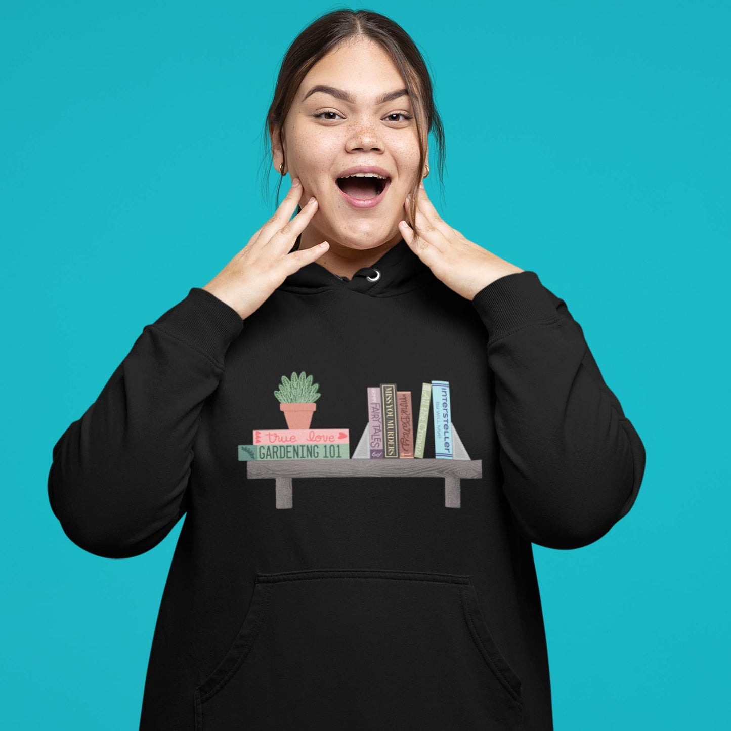 Moxie•Pop's unisex graphic hoodie with a illustration of a shelf, a series of books atop, and a house plant in black modeled by a woman with a turquoise backdrop