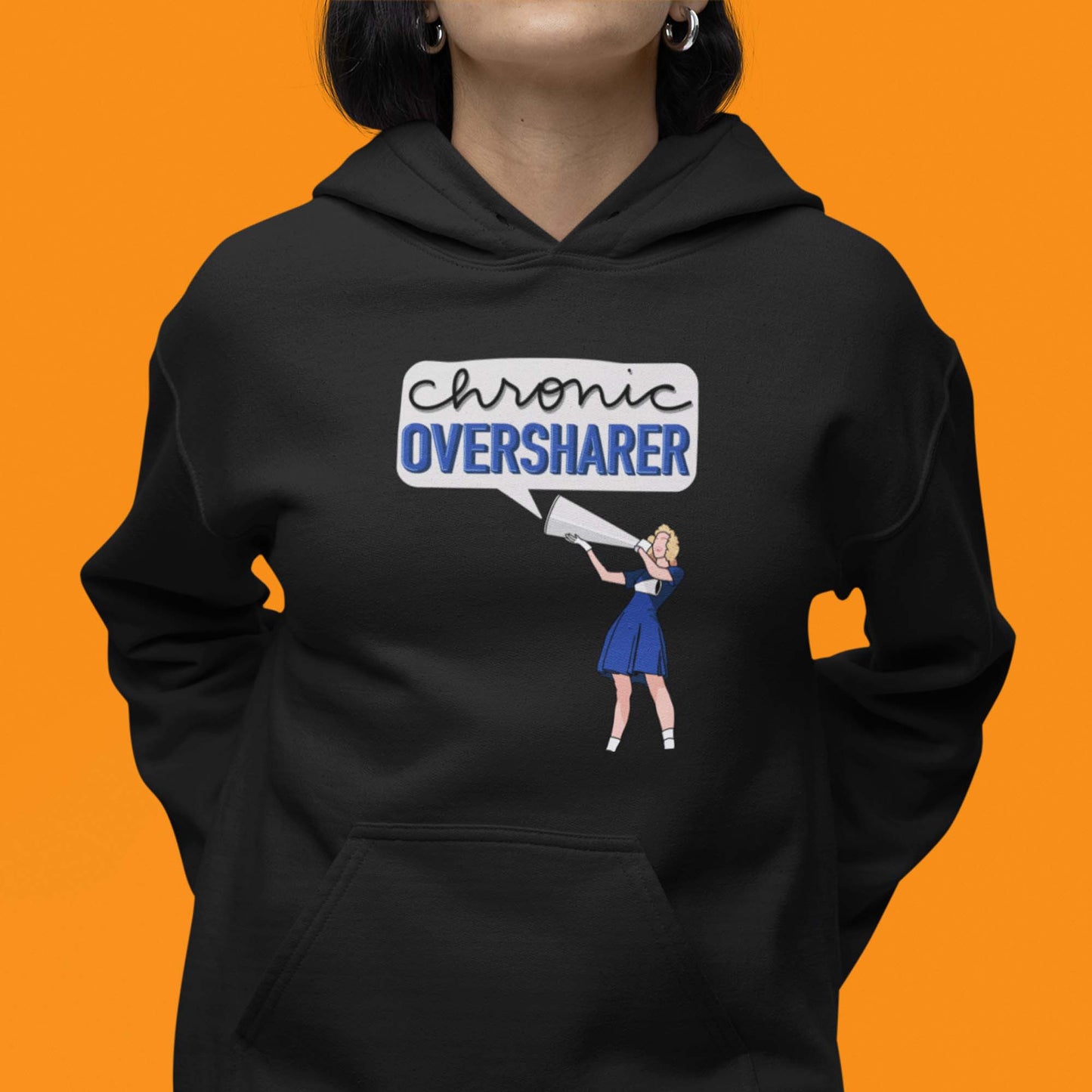 Moxie•Pop's unisex graphic hoodie with a illustration of a vintage cheerleader yelling into a megaphone with a talk bubble announcing 'Chronic Oversharer' in black modeled by a woman with an orange backdrop. 