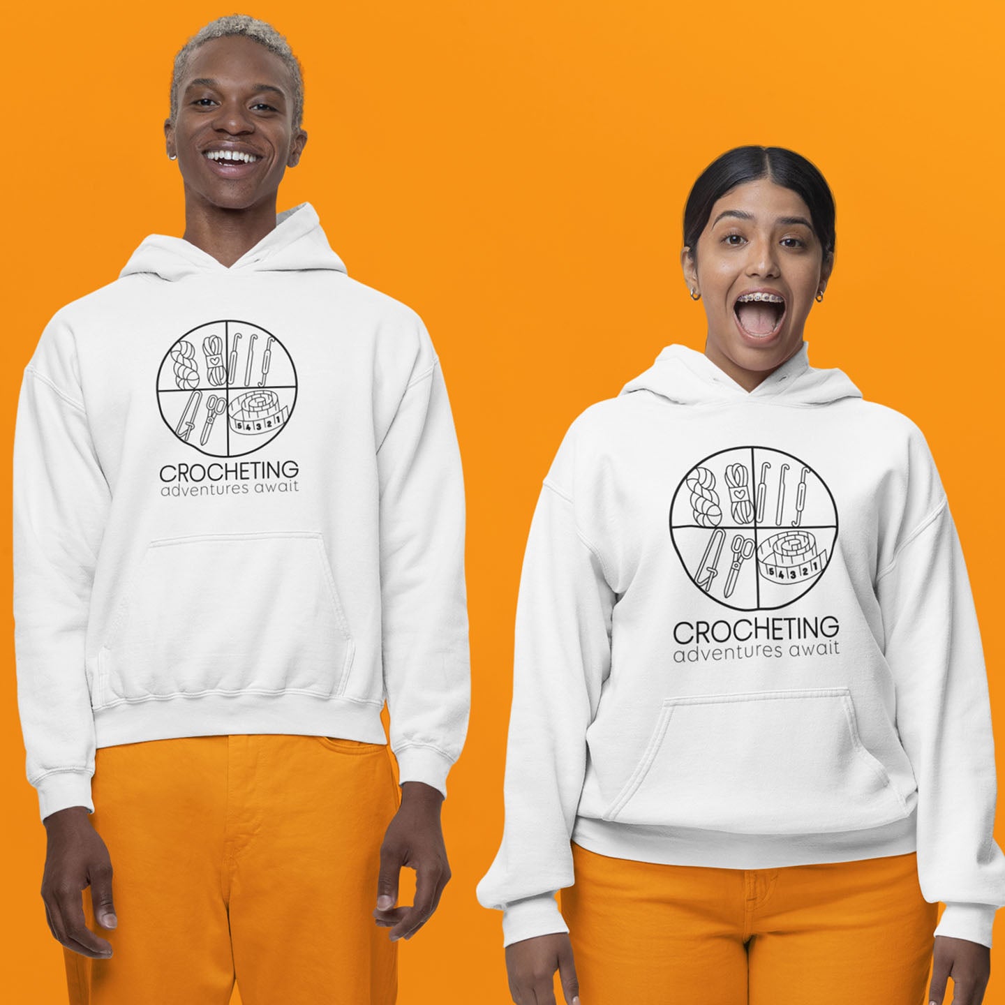 Modeled product image of Moxie•Pop's unisex graphic hoodie with "CROCHETING adventures await" below a circle filled with crocheting implements icons design in white. Models are wearing orange pants in front of a orange backdrop.