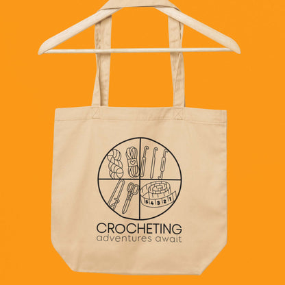 Hanging on a wood hanger product image of Moxie•Pop's eco tote bag with "CROCHETING adventures await" below a circle filled with crocheting implements icons design in oyster with an orange backdrop