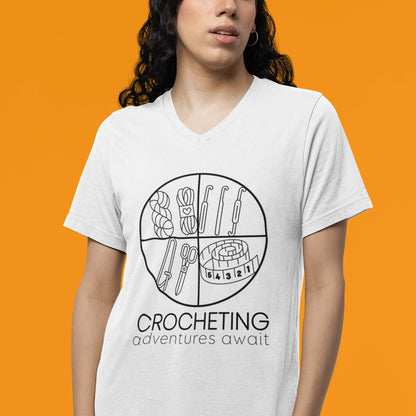 Moxie•Pop's unisex v-neck graphic tee with "CROCHETING adventures await" below a circle filled with crocheting implements icons design in white being modeled by a woman with an orange backdrop