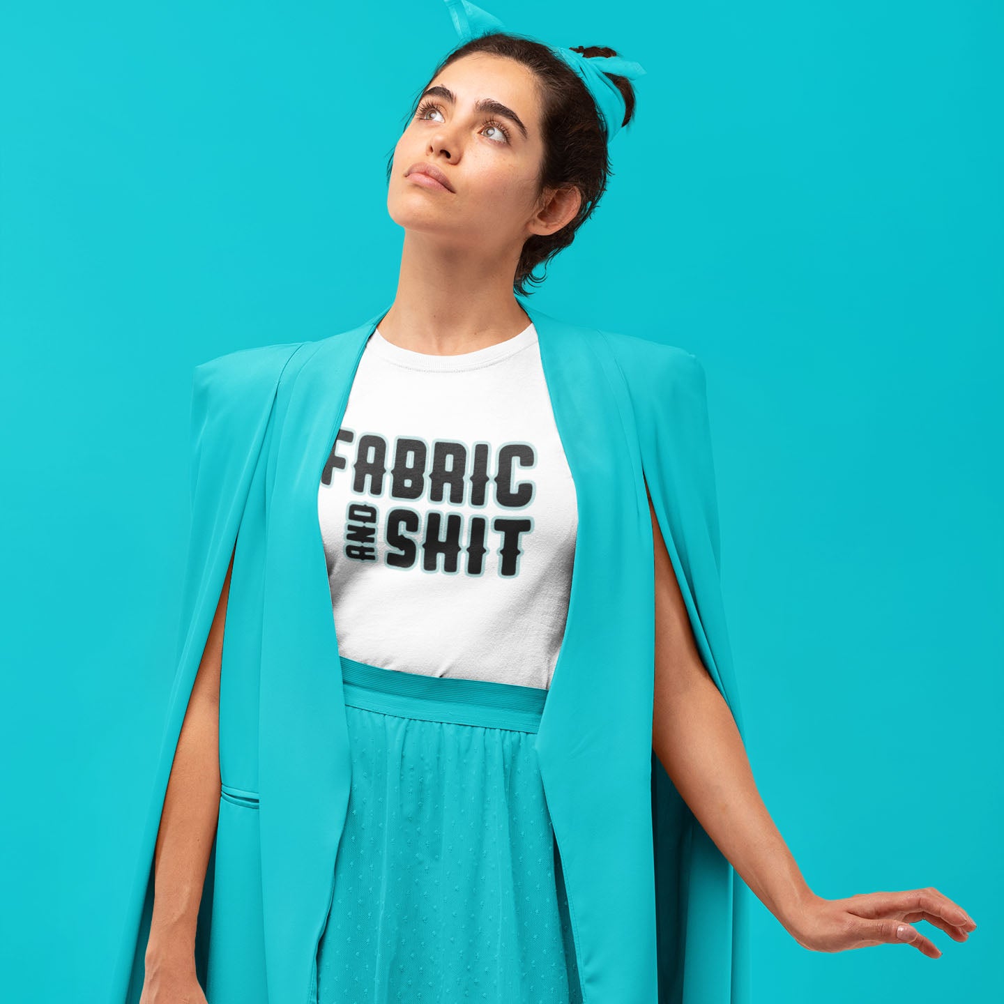 Modeled product image for Moxie•Pop's unisex graphic tee with "Fabric and Shit" typography design white. Model is wearing a turquoise cape and skirt in front of a turquoise backdrop. 