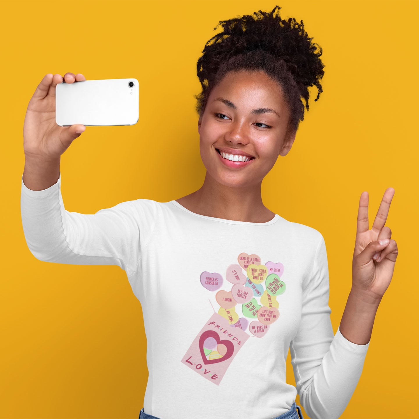 Modeled product image of Moxie•Pop's men's long sleeve graphic tee with Valentine's conversation hearts with quotes from the Friends TV show and a box that reads "Friends Love" in White. Model is taking a selfie while throwing a peace sign