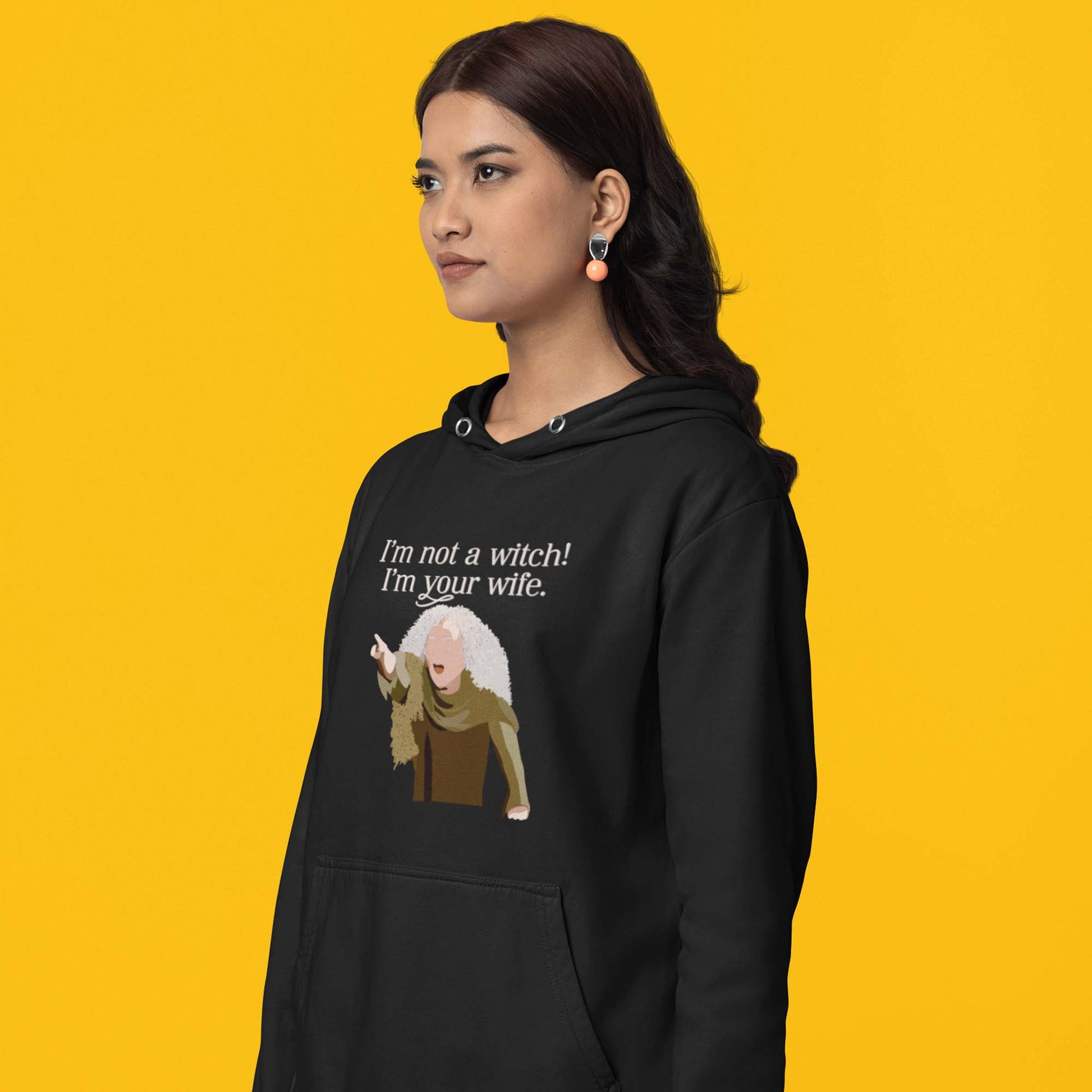 Moxie•Pop's unisex graphic hoodie with a illustration Valerie from The Princess Bride with the phrase 'I'm Not a Witch! I'm you wife." in black modeled by a woman with a yellow backdrop. 