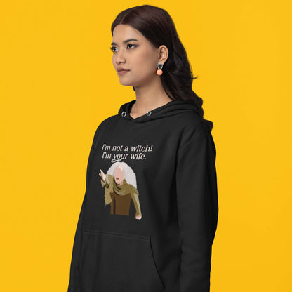 Moxie•Pop's unisex graphic hoodie with a illustration Valerie from The Princess Bride with the phrase 'I'm Not a Witch! I'm you wife." in black modeled by a woman with a yellow backdrop. 