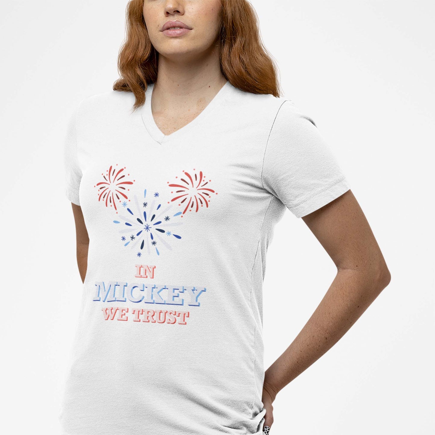 Moxie•Pop's unisex v-neck graphic tee with hand-drawn fireworks in the shape of Mickey Mouse silhouette with the phrase "In Mickey We Trust" in white modeled by a woman with orange hair with with a white backdrop. 