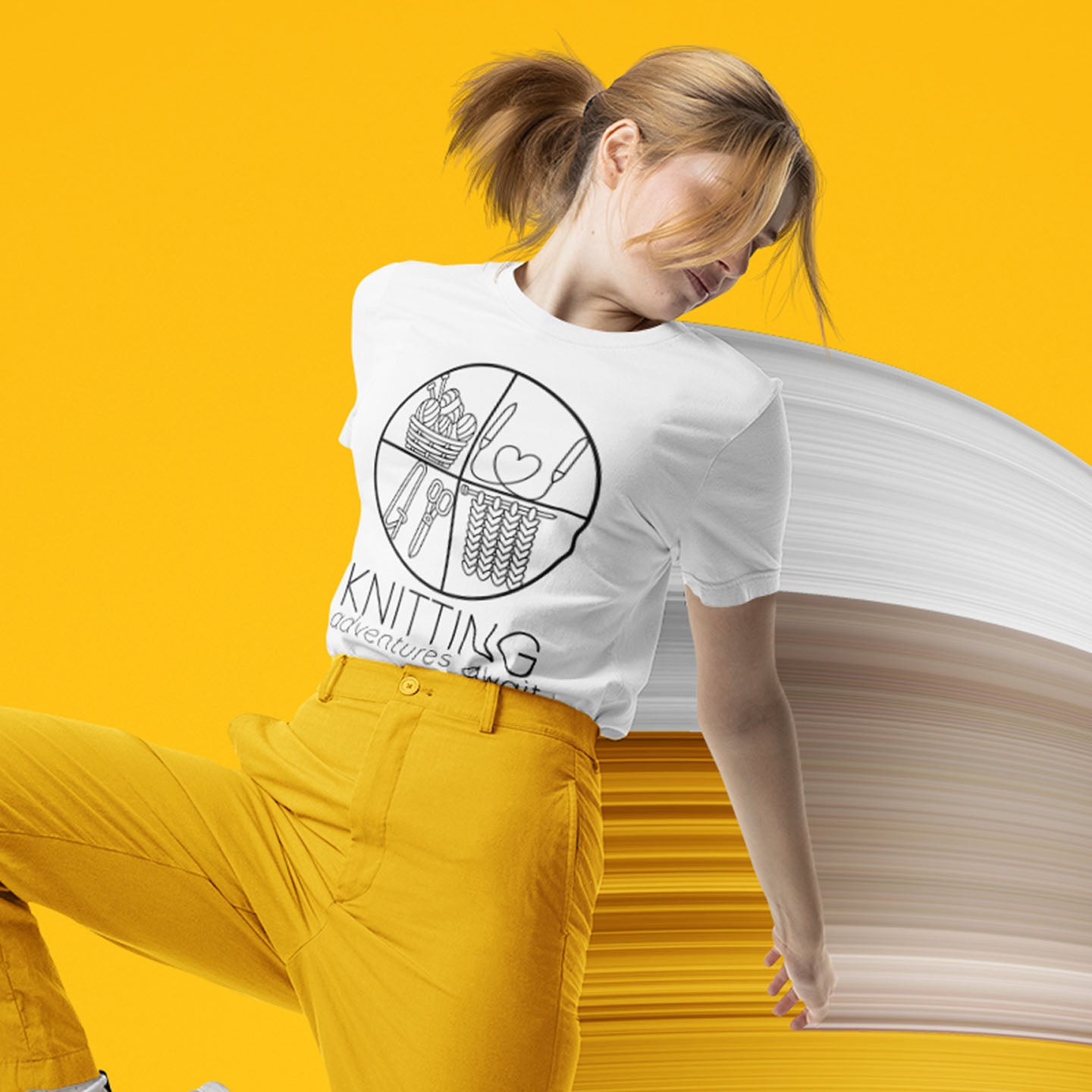 Modeled product image of Moxie•Pop's unisex graphic tee with "KNITTING adventures await" below a circle filled with knitting implements icons design in white. Model is wearing yellow pants 