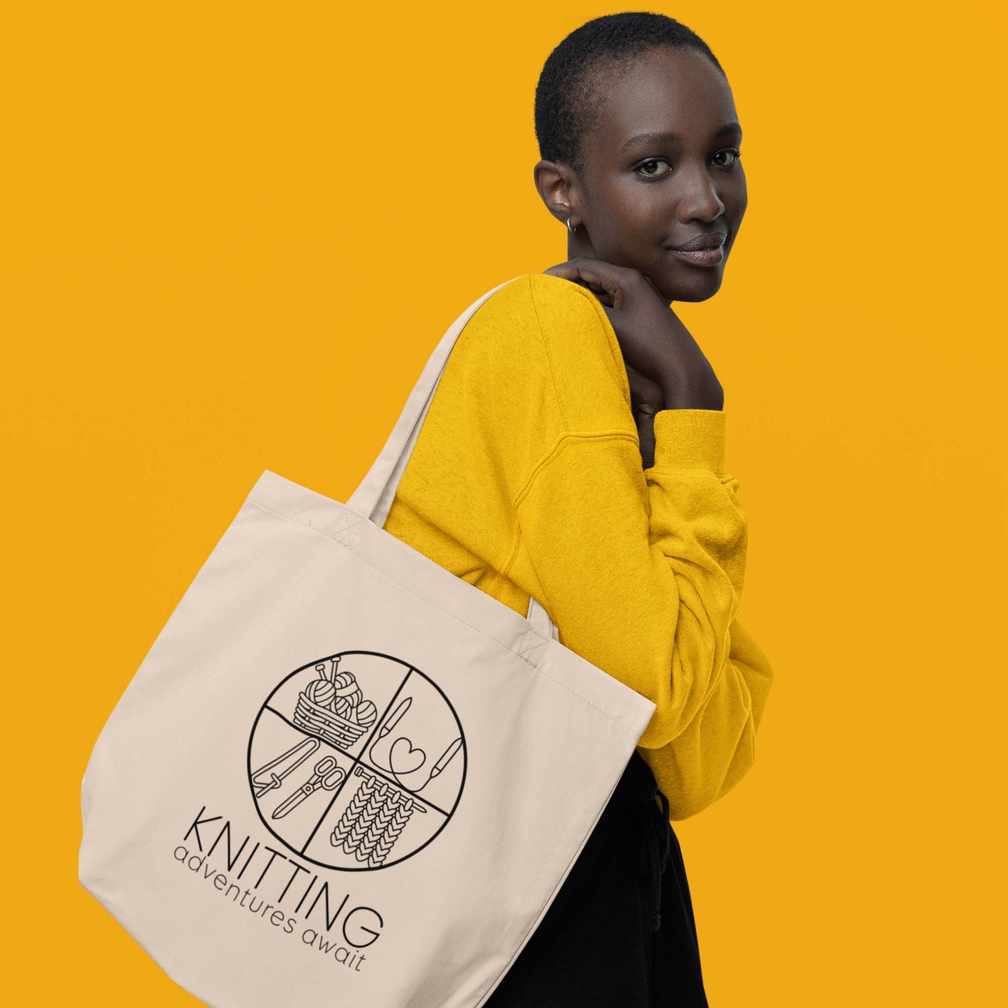 Moxie•Pop's eco tote bag with "KNITTING adventures await" below a circle filled with knitting implements icons design in oyster model by a woman with short hair in a yellow sweatshirt with yellow backdrop. 