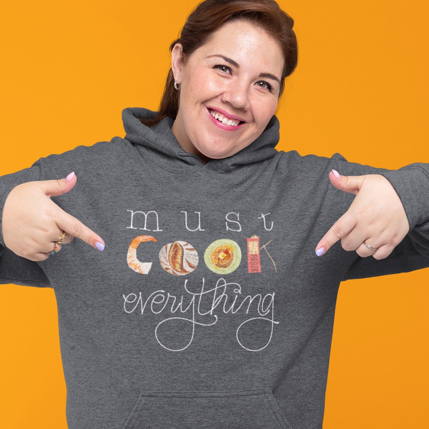 Modeled product image of Moxie•Pop's unisex graphic hoodie with hand lettered 'must cook everything' with the word cook created from hand drawn food items in dark grey heather