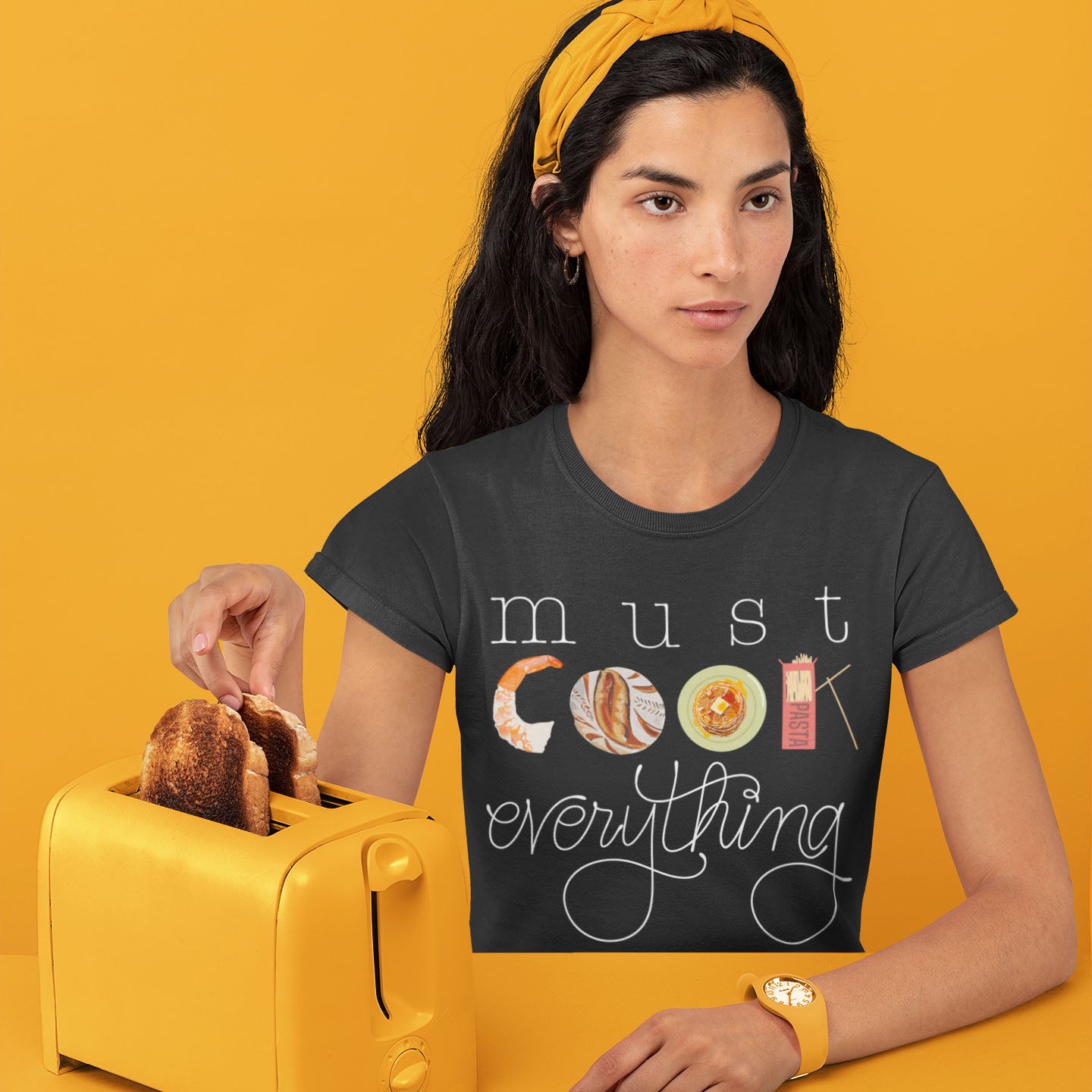 Modeled product image of Moxie•Pop's unisex graphic tee with hand lettered 'must cook everything' with the word cook created from hand drawn food items in dark grey heather. Model is sitting in front of a orange backdrop at an orange table, with an orange toaster, wearing and orange watch. 