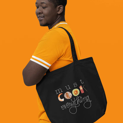 Moxie•Pop's eco tote bag with hand lettered 'must cook everything' with the word cook created from hand drawn food items in black modeled by man in an orange shirt with an orange backdrop