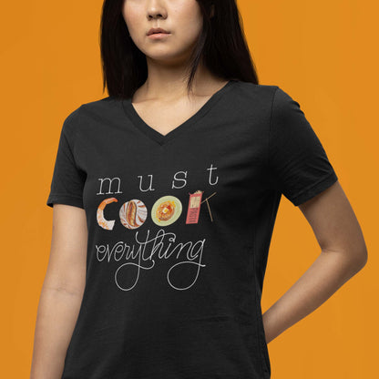 Moxie•Pop's unisex v-neck graphic tee with hand lettered 'must cook everything' with the word cook created from hand drawn food items in black modeled by a woman with long hair and an orange backdrop