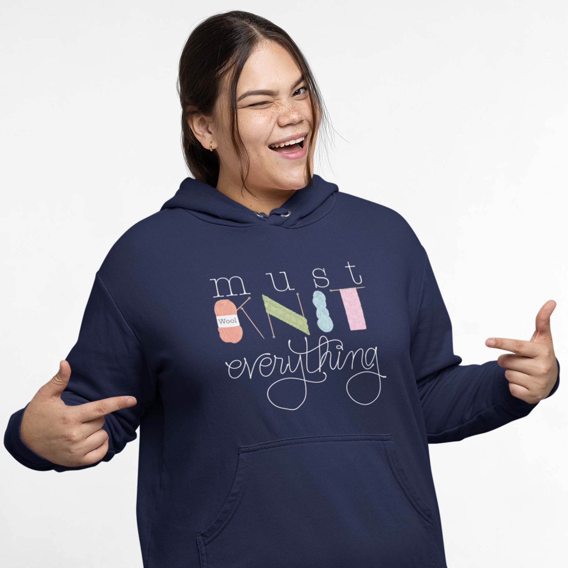 Moxie•Pop's unisex graphic hoodie with hand lettered 'Must Knit Everything' with the word knit created from illustrations of yarn, needles, and knitting in navy modeled by a woman with a pony tail and a white backdrop