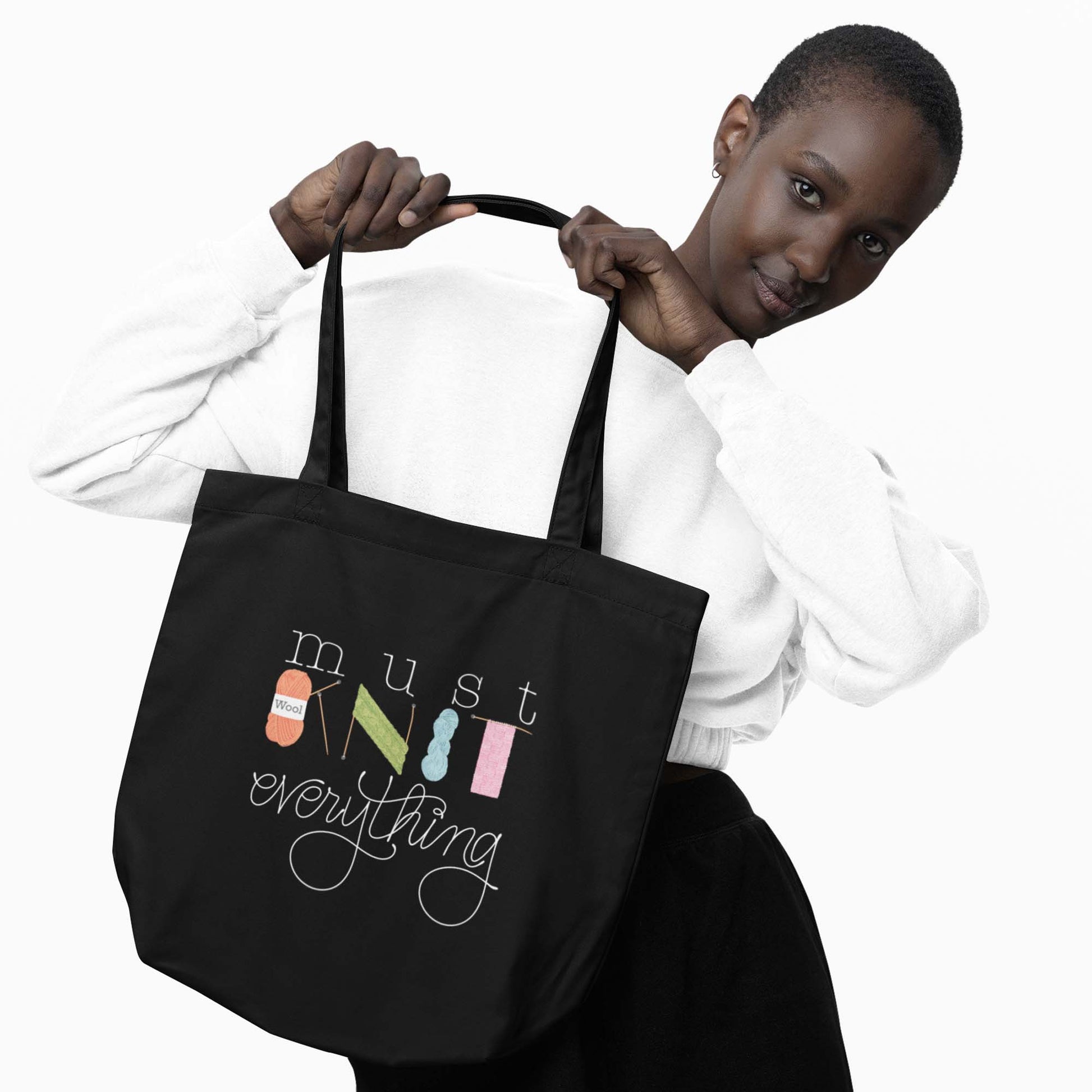 Moxie•Pop's eco tote bag with hand lettered 'Must Knit Everything' with the word knit created from illustrations of yarn, needles, and knitting in black being held by a woman with short hair