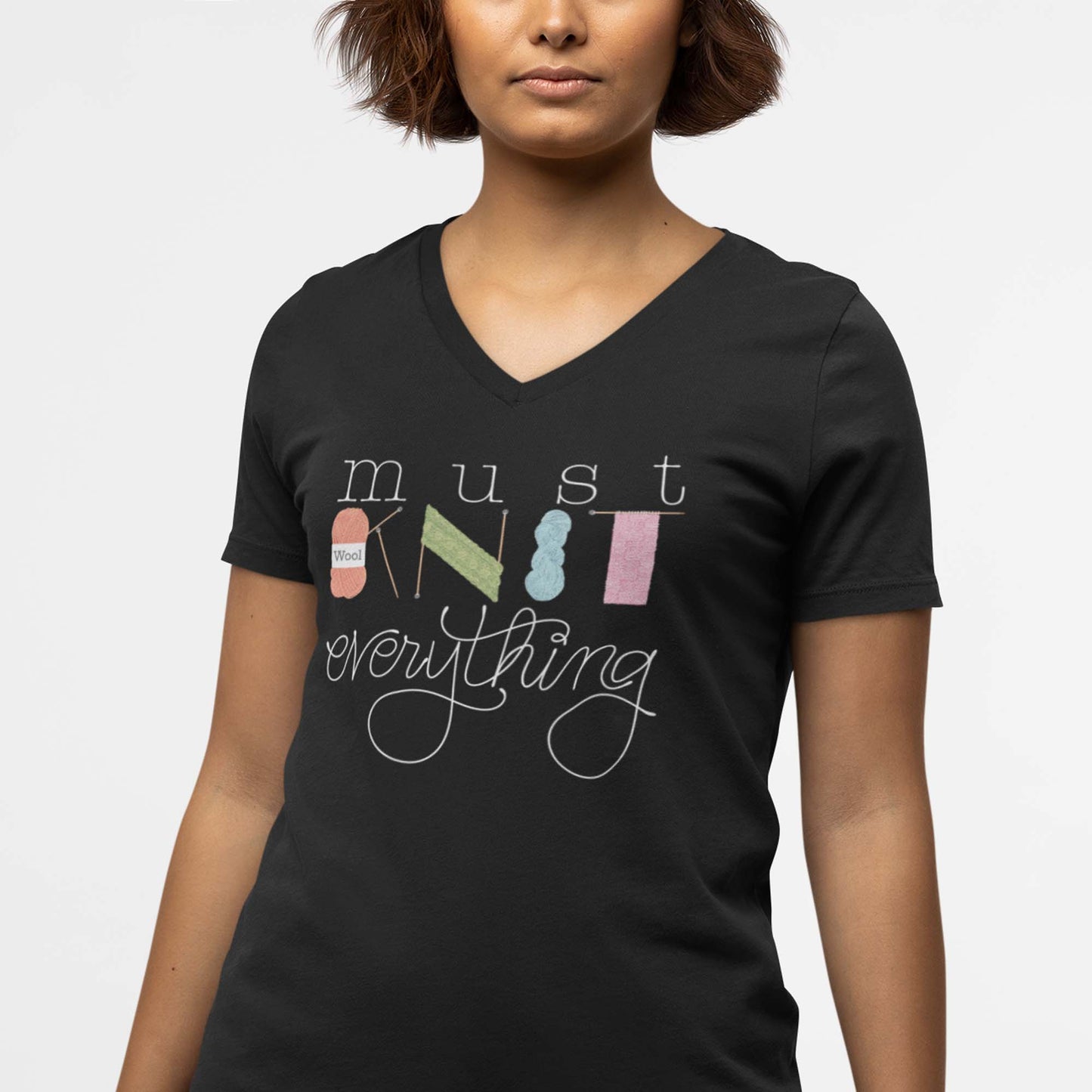 Moxie•Pop's unisex v-neck tee with hand lettered "must KNIT everything" with the downstrokes of the word 'KNIT' hand drawn yarn, knitting needles, and knitted scarves modeled by a woman with short hair and a white backdrop. 