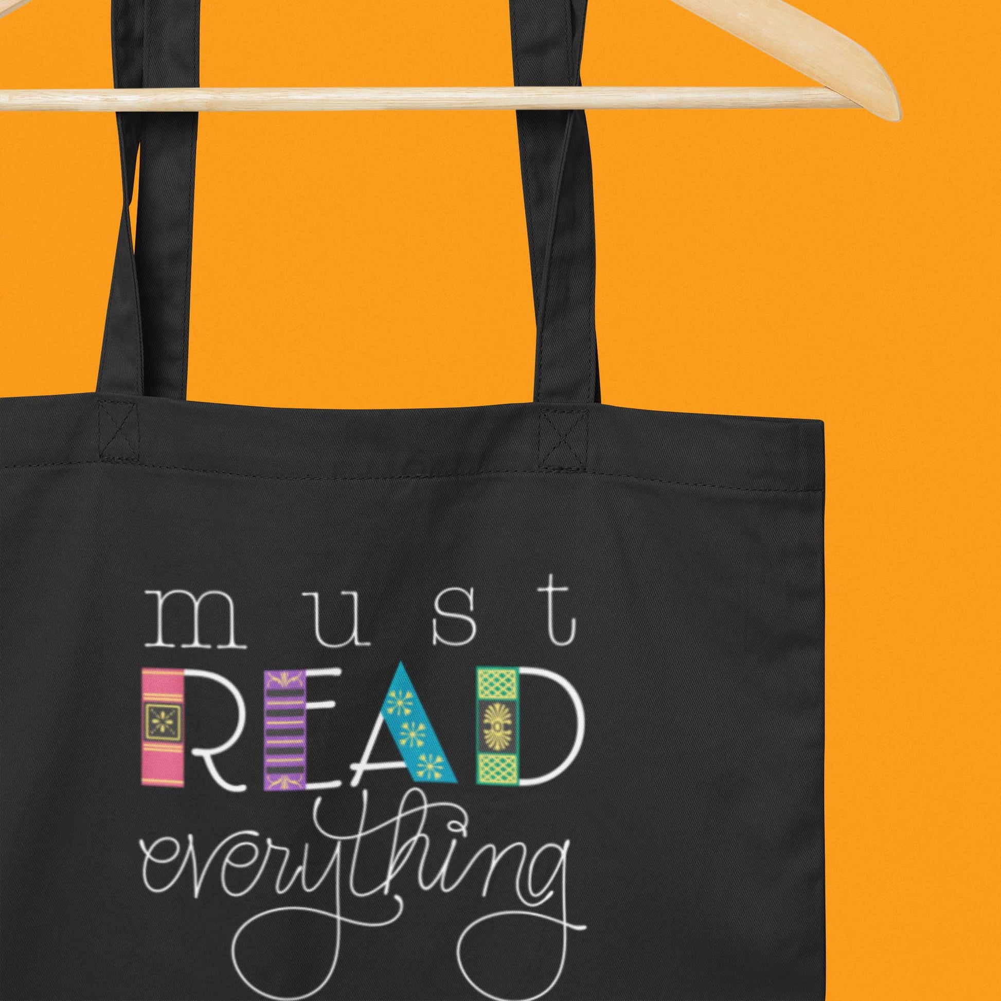 Hanging product image of Moxie•Pop's eco tote bag with hand lettered "must READ everything" with the downstrokes of the word 'read' drawn to look like book spines in black with an orange backdrop. 