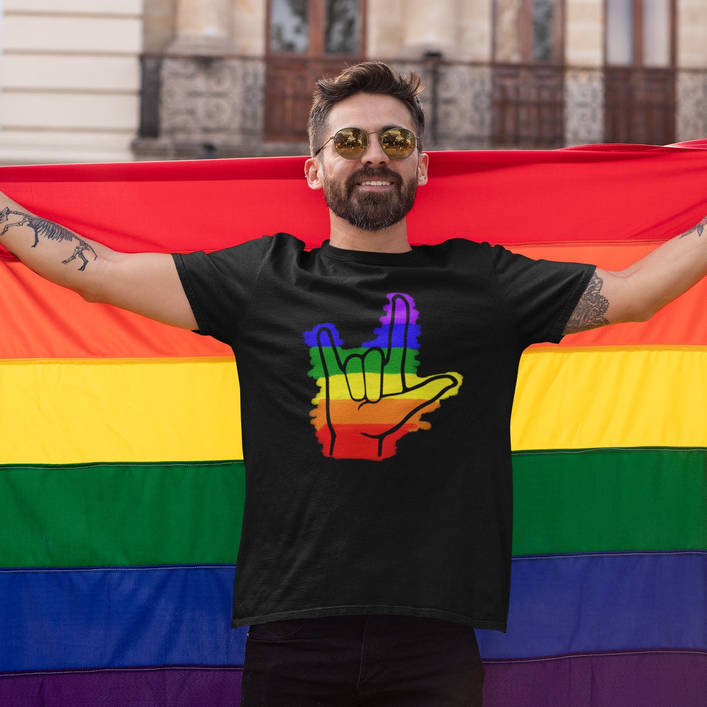 Modeled product image of Moxie•Pop's unisex graphic tee with colored rainbow stripes behind a black ASL I love you hand sign icon design in black heather. Modeled is holding a pride flag.