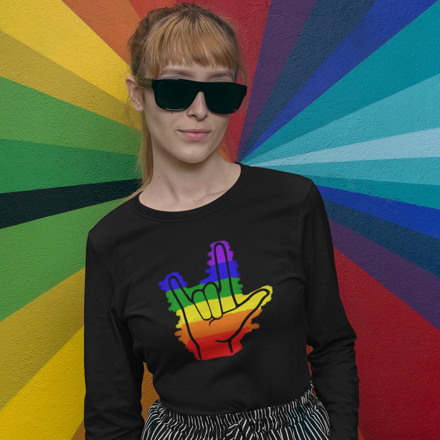 Modeled product image of Moxie•Pop's men's long sleeve graphic tee with colored rainbow stripes behind a black ASL I love you hand sign icon design in black. Model is wearing black sunglasses and is standing front of a rainbow colored wall. 