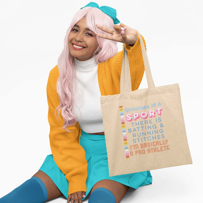 Moxie•Pop's eco tote bag  in oyster with "Quilting is a sport. There's batting and running stitches. I'm basically a pro athlete" graphic being held by a woman sitting with a long pink hair wearing a yellow sweater, a turquoise skirt and bow with a white backdrop. 