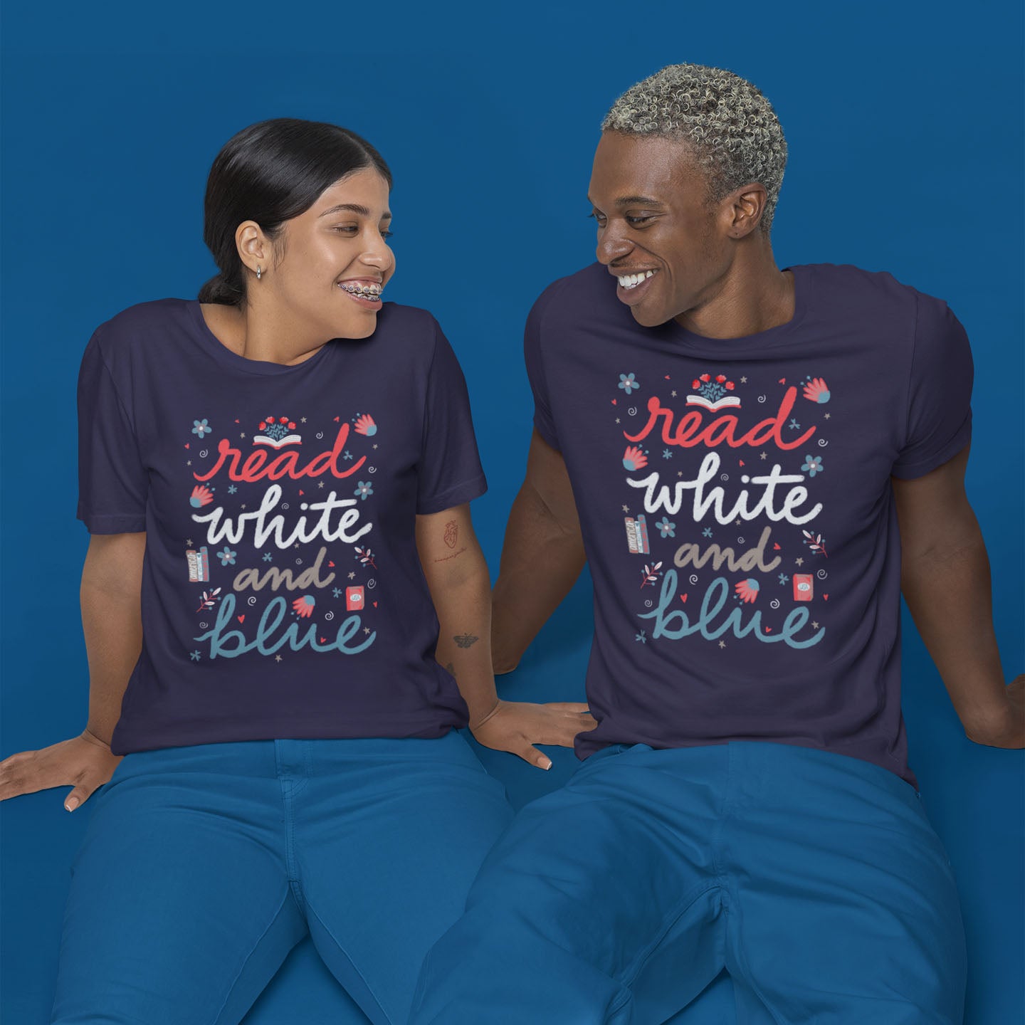 Modeled product image of Moxie•Pop's unisex graphic tee with "read white and blue" surrounded by hand-drawn flowers and books design in heather midnight navy. Models are sitting on the floor in front of a blue backdrop wearing blue pants. 