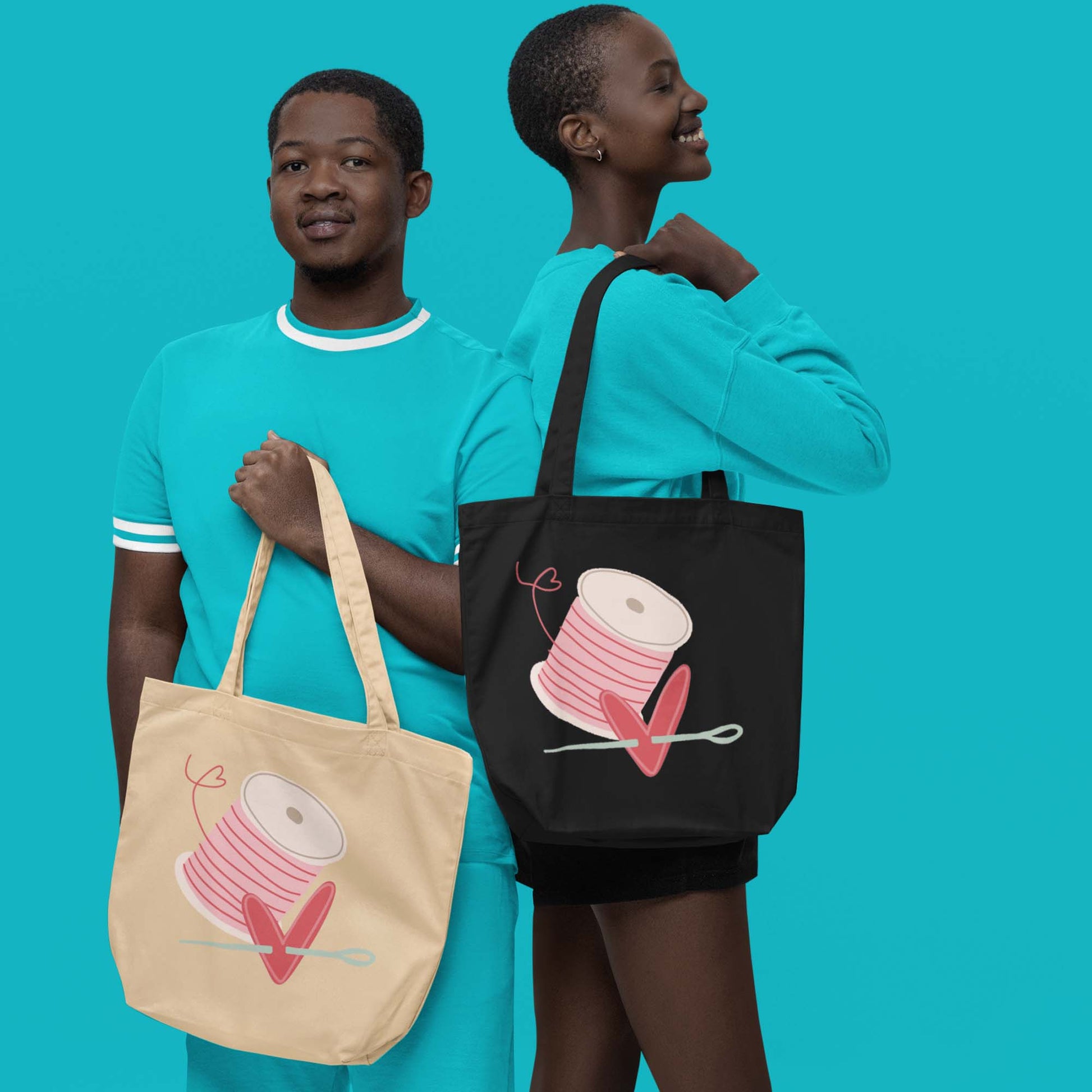 Moxie•Pop's eco tote bag in oyster with a spool of pink thread and a heart pierced by a needle hand-drawn design in oyster and black modeled by a man and a woman wearing turquoise with a turquoise backdrop. 