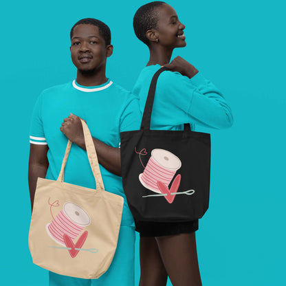 Moxie•Pop's eco tote bag in oyster with a spool of pink thread and a heart pierced by a needle hand-drawn design in oyster and black modeled by a man and a woman wearing turquoise with a turquoise backdrop. 