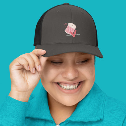 Moxie•Pop's retro trucker hat in charcoal with black mesh embroidered with pink spool of thread with a heart being pierced by a needle modeled by a woman in a turquoise sweater and a turquoise backdrop. 