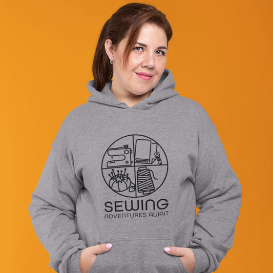 Modeled product image Moxie•Pop's unisex graphic hoodie with a black circle outline in four quadrants each with a sewing tool icon above 'Sewing Adventures Await' in sport grey