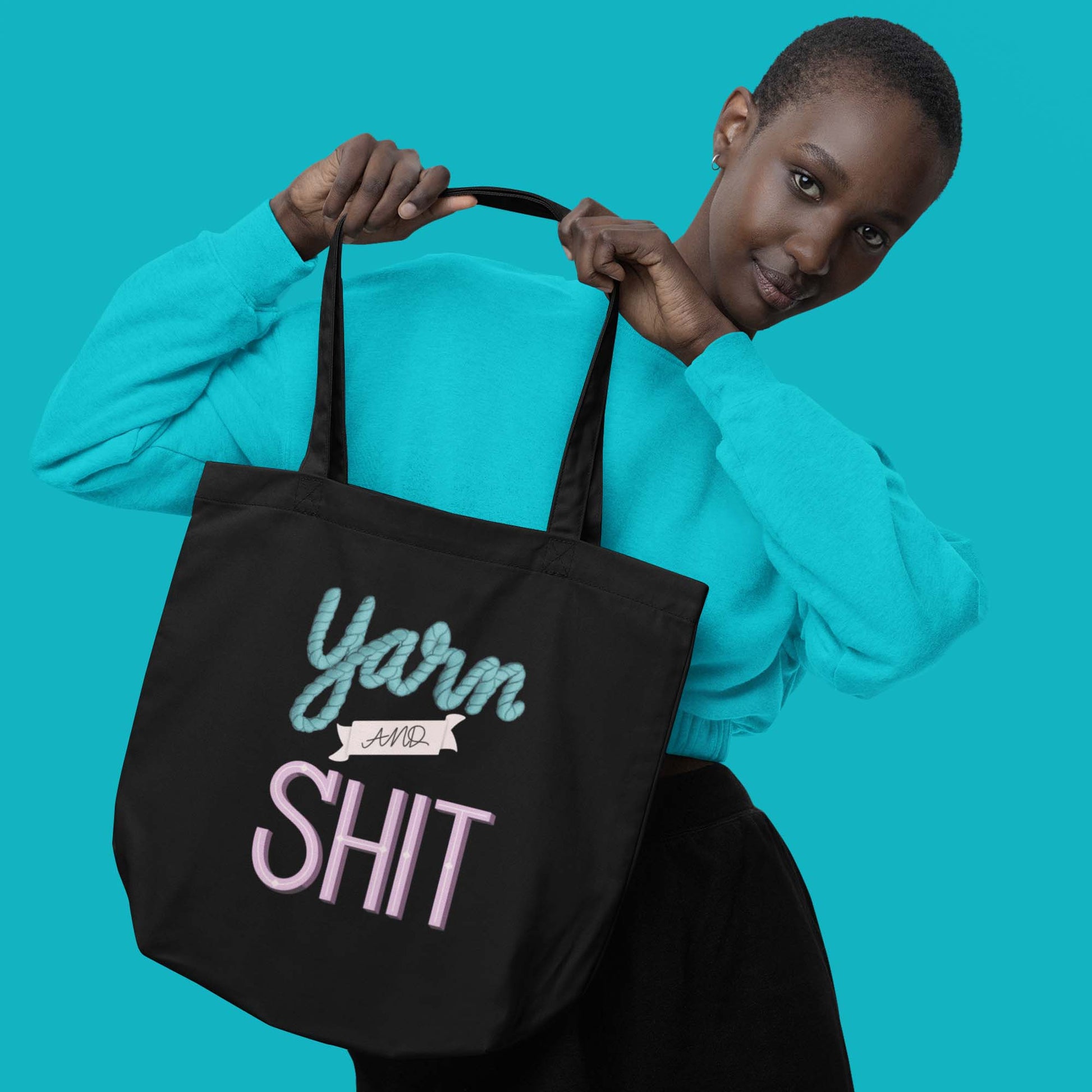 Moxie•Pop's eco tote bag with hand lettered "Yarn and Shit" design in black modeled by a woman with short hair in turquoise sweatshirt with turquoise backdrop. 