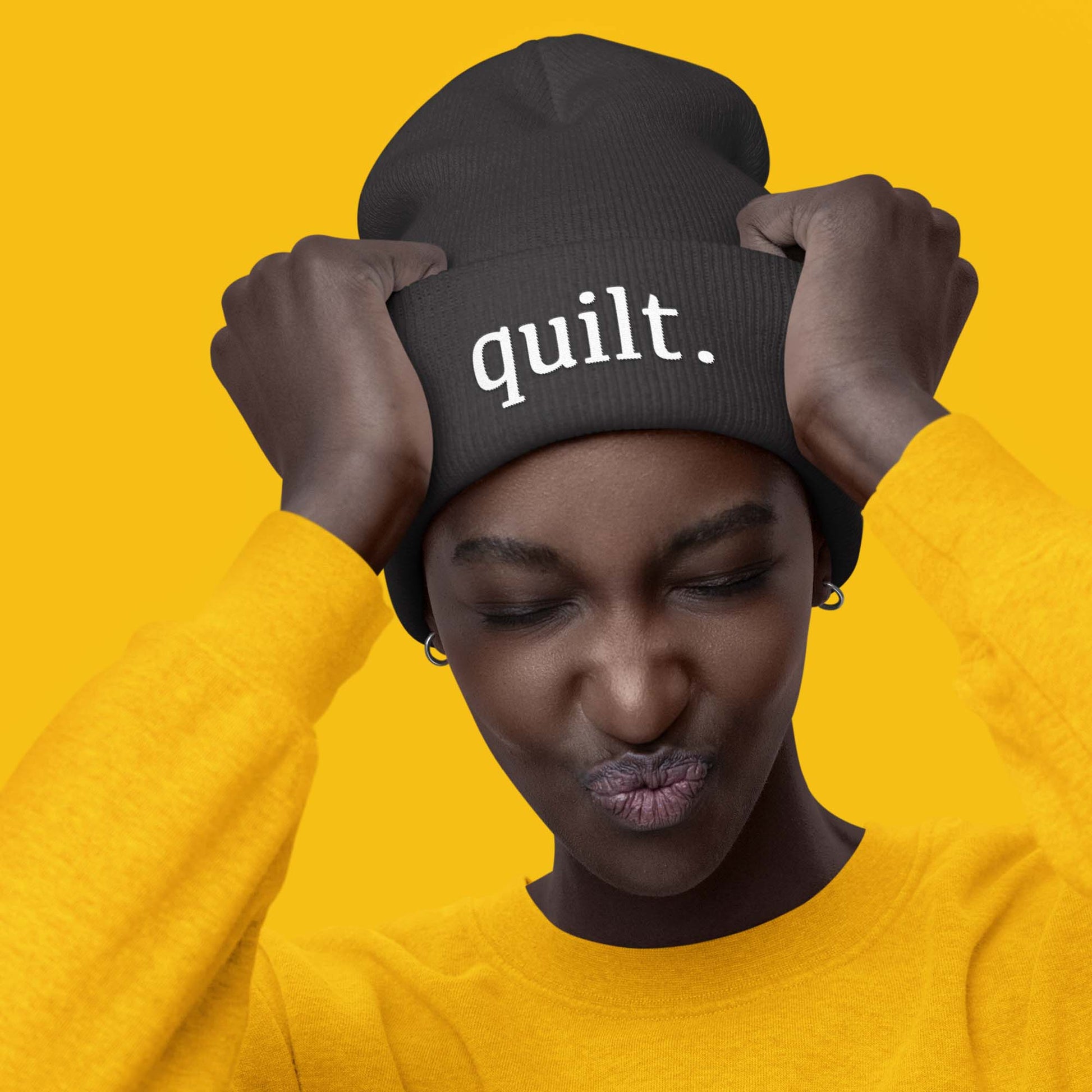 Moxie•Pop's cuffed beanie in dark grey with "quilt." embroidered in white modeled by a woman wearing a yellow sweatshirt and a yellow backdrop. 