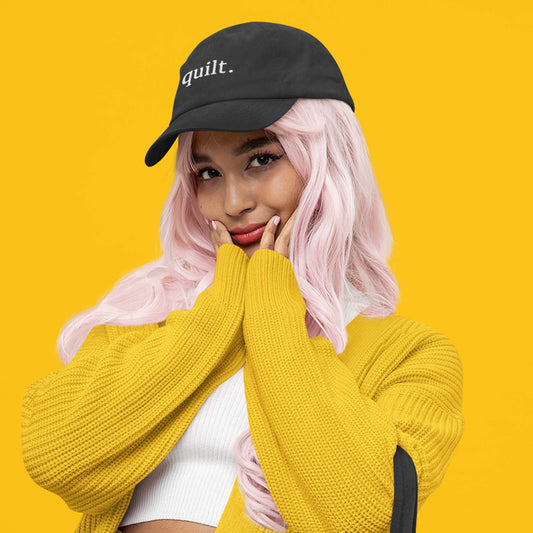 Moxie•Pop's embroidered dad hat with the word "quilt." in black modeled by a woman with long pink hair wearing a yellow sweater and a yellow backdrop