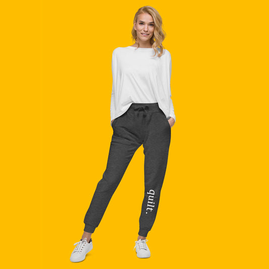 Moxie•Pop's unisex fleece jogger sweatpants with "quilt." on the front left shin in charcoal heather modeled by a woman in a white shirt and tennis shoes with a yellow backdrop