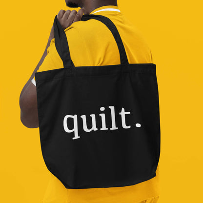 Moxie•Pop's eco graphic tote bag with 'quilt.' serif typography in black modeled by a man in a yellow shirt with a yellow backdrop.