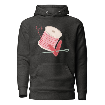 Moxie•Pop's unisex graphic hoodie in charcoal grey with a spool of pink thread and a heart pierced by a needle hand-drawn design.