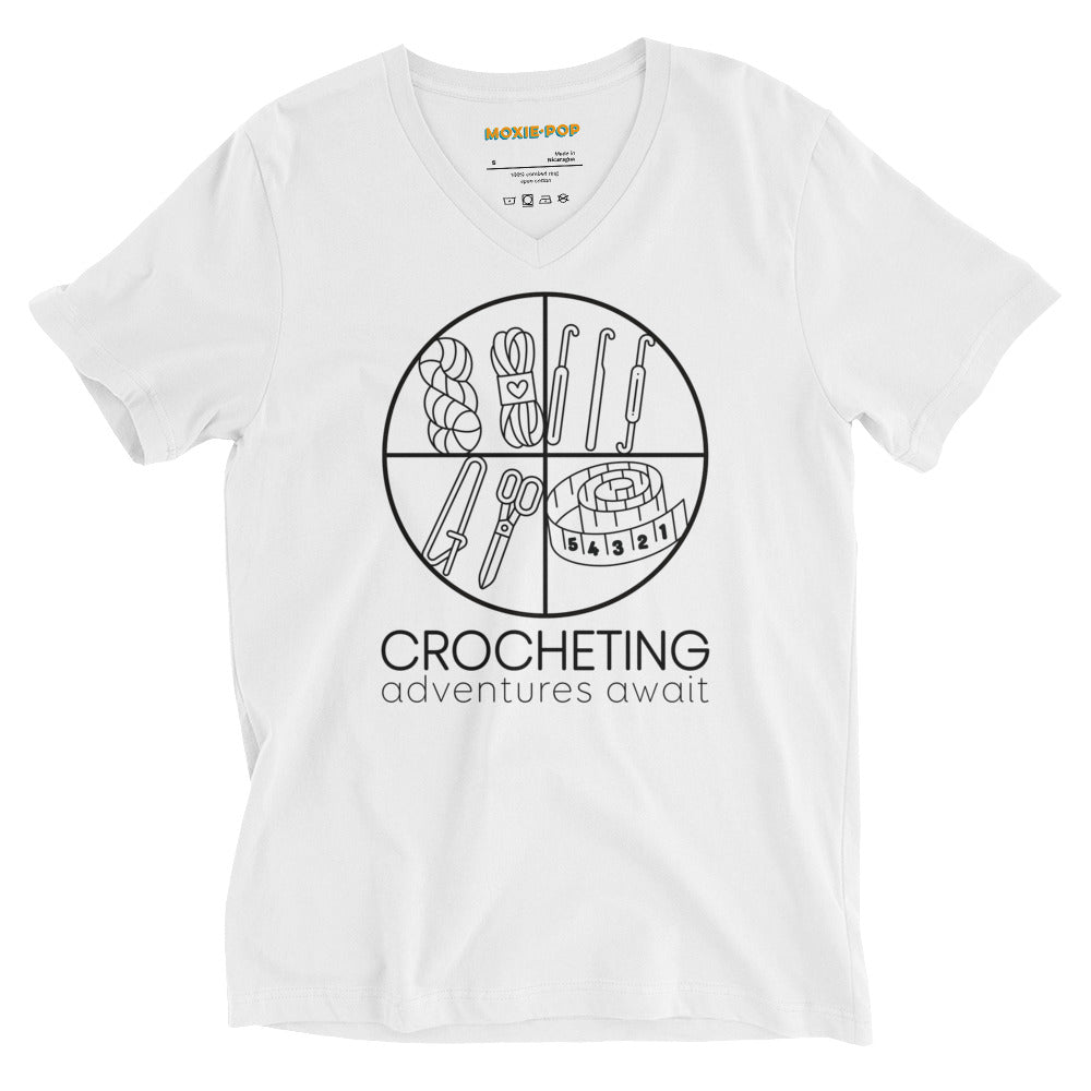 Lay flat product image of Moxie•Pop's unisex v-neck graphic tee with "CROCHETING adventures await" below a circle filled with crocheting implements icons design in white